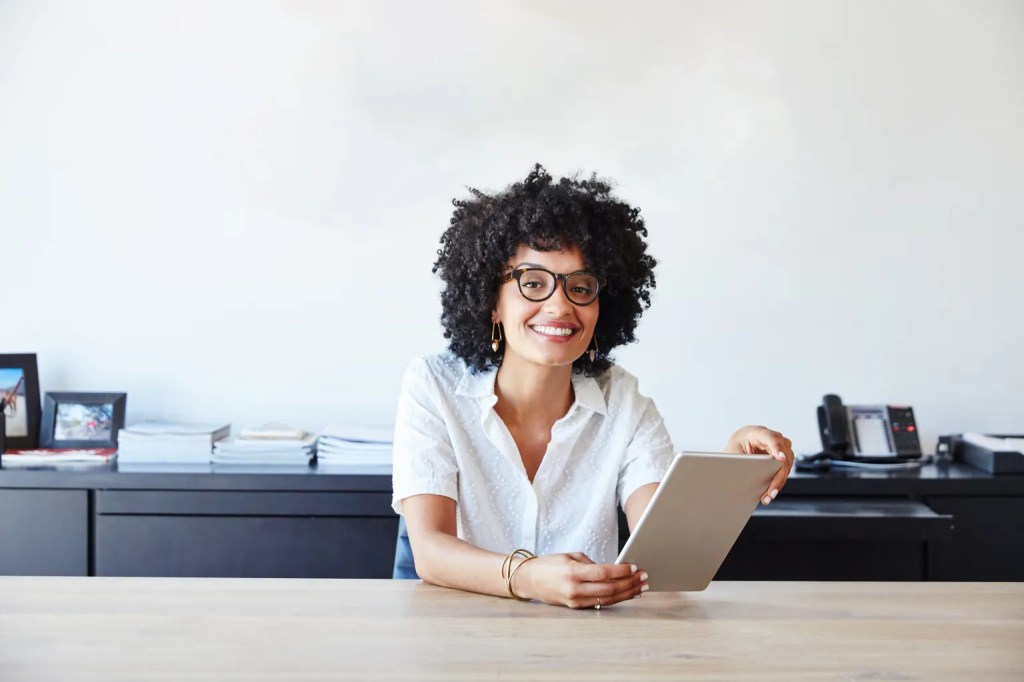 Woman with curly hair and glasses smiling while holding a tablet at her desk.