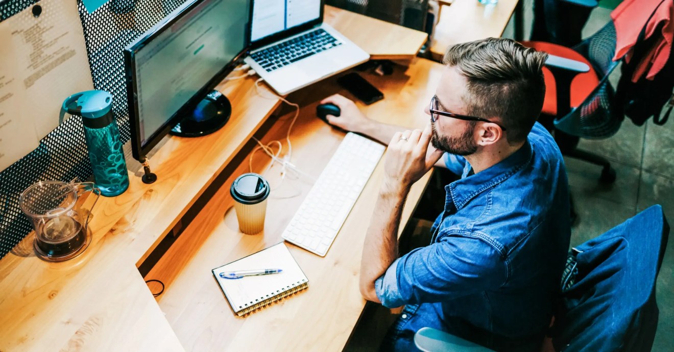 A person resting their hand on their chin while they work from a laptop connected to a monitor on a wooden desk