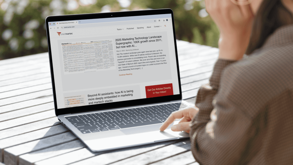 Woman sitting at an outdoor table using a laptop, browsing the Chief Martec website. The screen displays an article titled “2025 Marketing Technology Landscape Supergraphic: 100X growth since 2011, but now with AI…” with additional headlines and a red button prompting readers to subscribe via email.