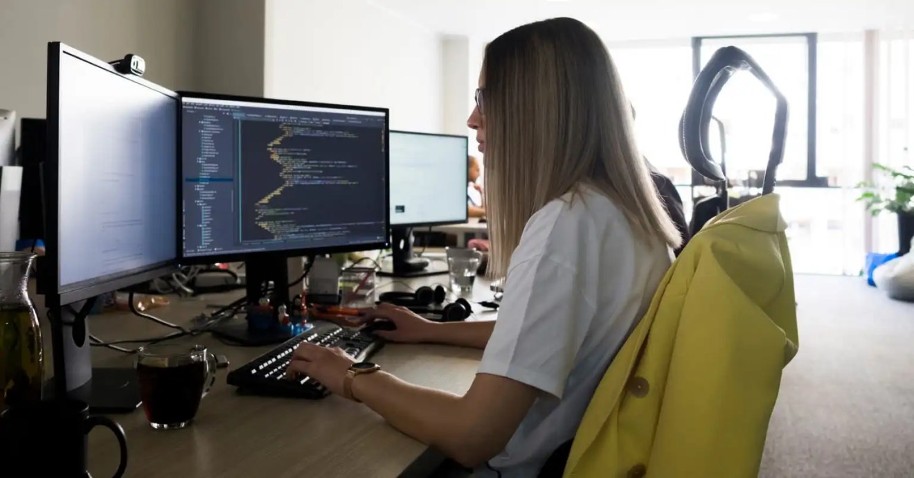 Woman coding at dual monitors, managing digital solution content workflow in a modern workspace.