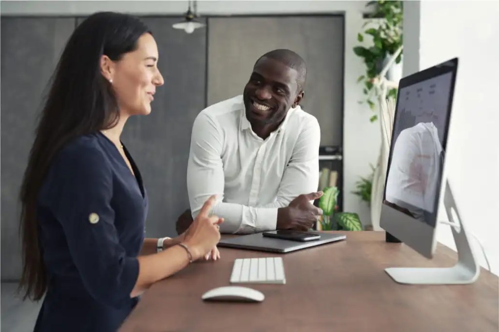 Two colleagues smiling and talking at a desk with a computer and tablet in a modern office, discussing wordpress vip premier support solutions.
