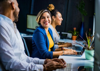 An image of a three-person team working from the same desk with two of them smiling at one another
