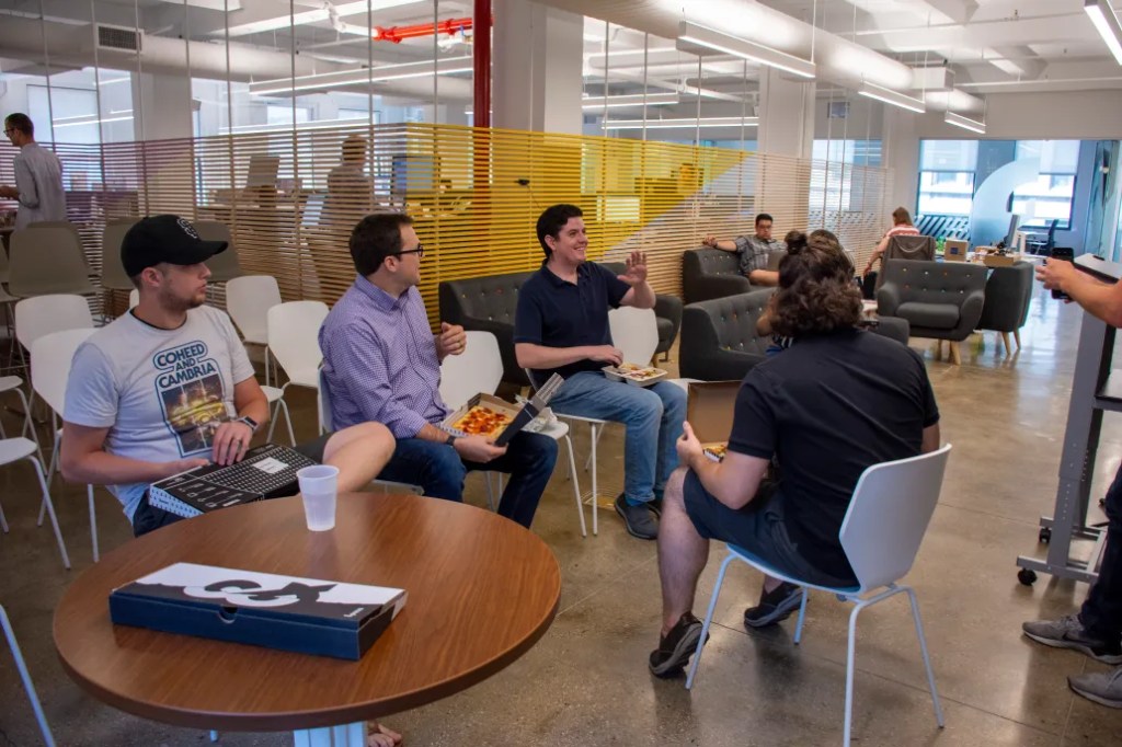 Group of people talking and eating lunch together in an open floor plan office space.