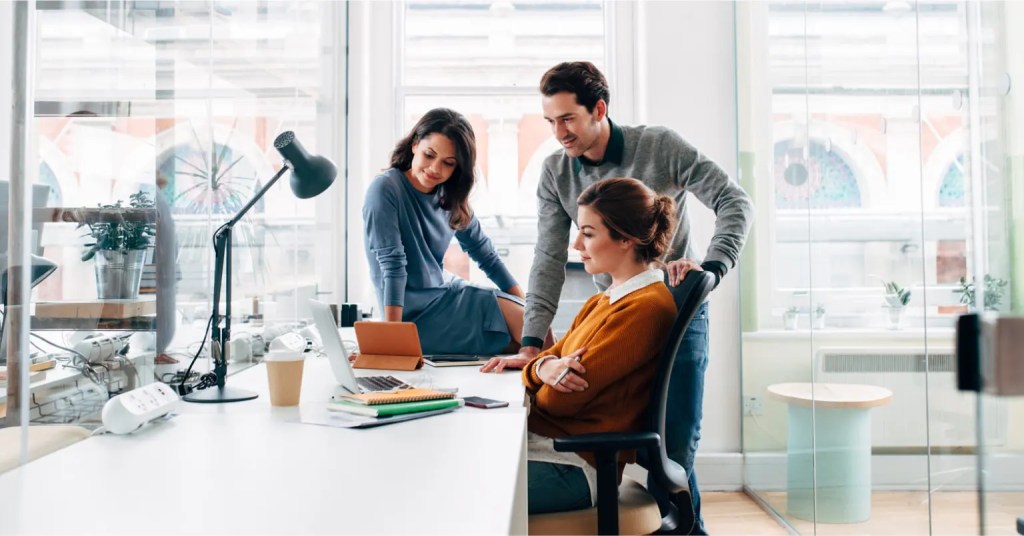 Three coworkers collaborating at a desk on digital solution content workflow using laptops.