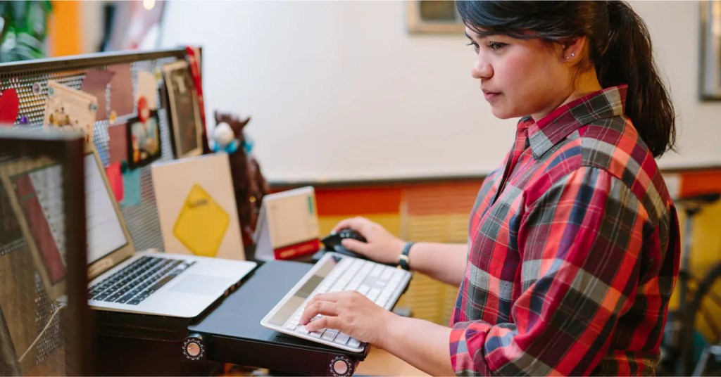Woman uses standing desk to manage content on digital solution editorial dashboard with laptop and monitor.