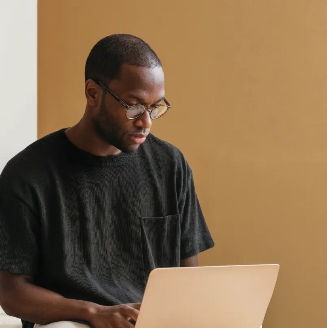 A person wearing glasses and a black T-shirt using a laptop while seated against a plain tan wall.
