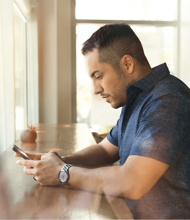 Person sitting at a wooden counter looking at a smartphone near a window.