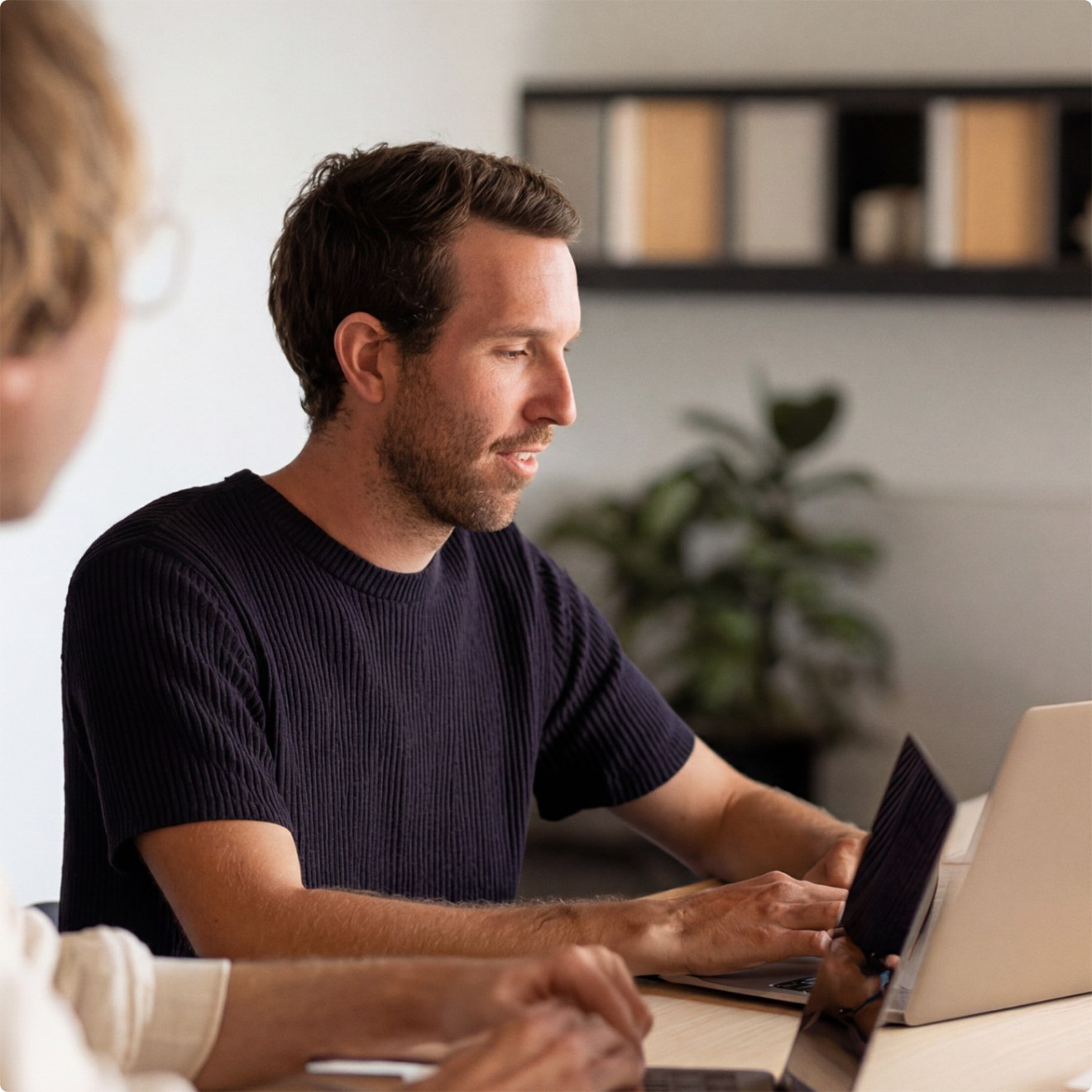 Two people working at laptops in a bright office, with shelves and a plant in the background.