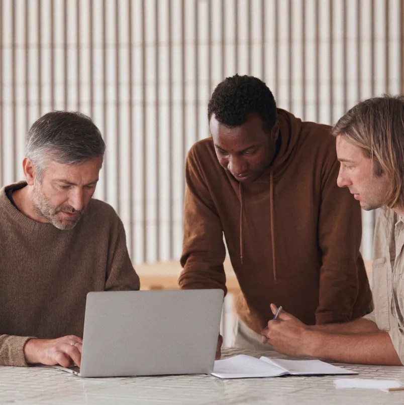 Three colleagues collaborating around a laptop while reviewing notes and discussing a project.