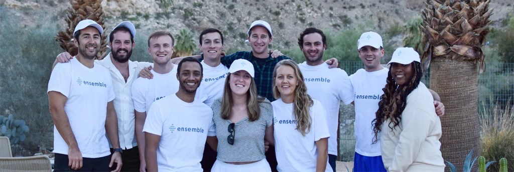 A group of eleven people smiling and posing outdoors in front of palm trees and desert hills. Most are wearing white “ensemble” t-shirts and white caps, suggesting a team gathering or company event.
