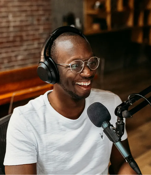 Person wearing glasses and over-ear headphones, smiling while speaking into a microphone mounted on a boom arm in a podcast or recording studio setting.