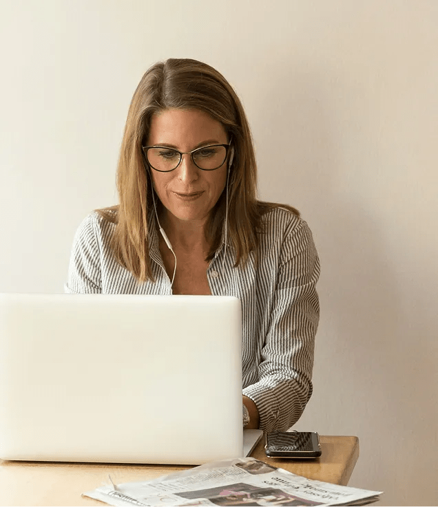 A professional wearing glasses and earbuds working on a laptop at a desk, with a smartphone and printed documents nearby.