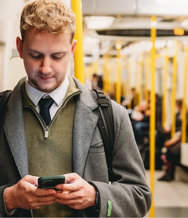 A commuter standing inside a train, looking at a smartphone while wearing business attire and a backpack.