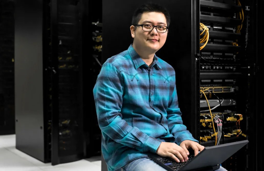 Man smiling at camera sitting near servers with a laptop
