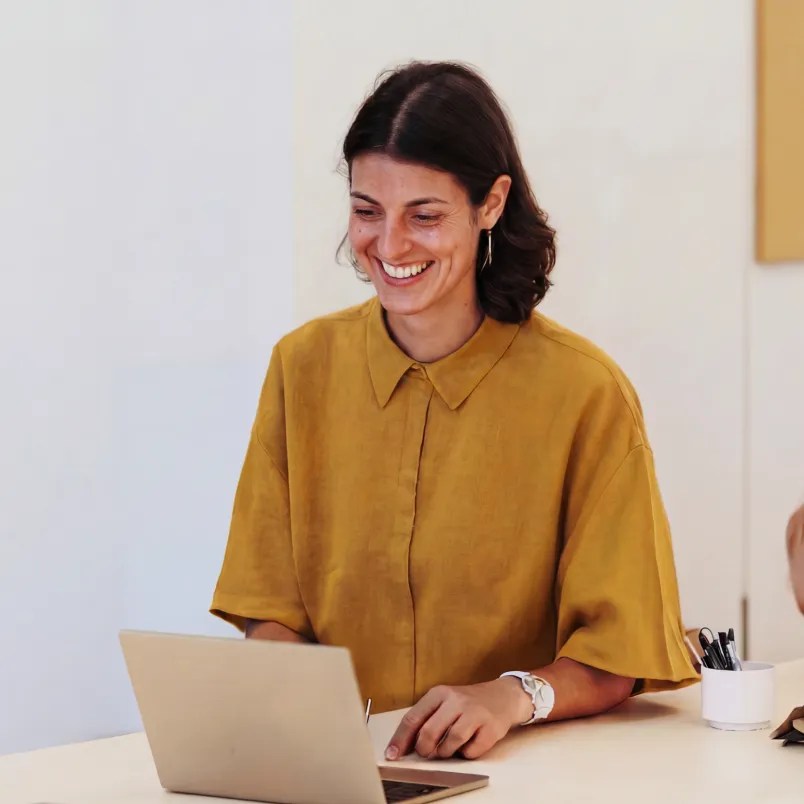 Person smiling while working on a laptop at a desk in a bright office.