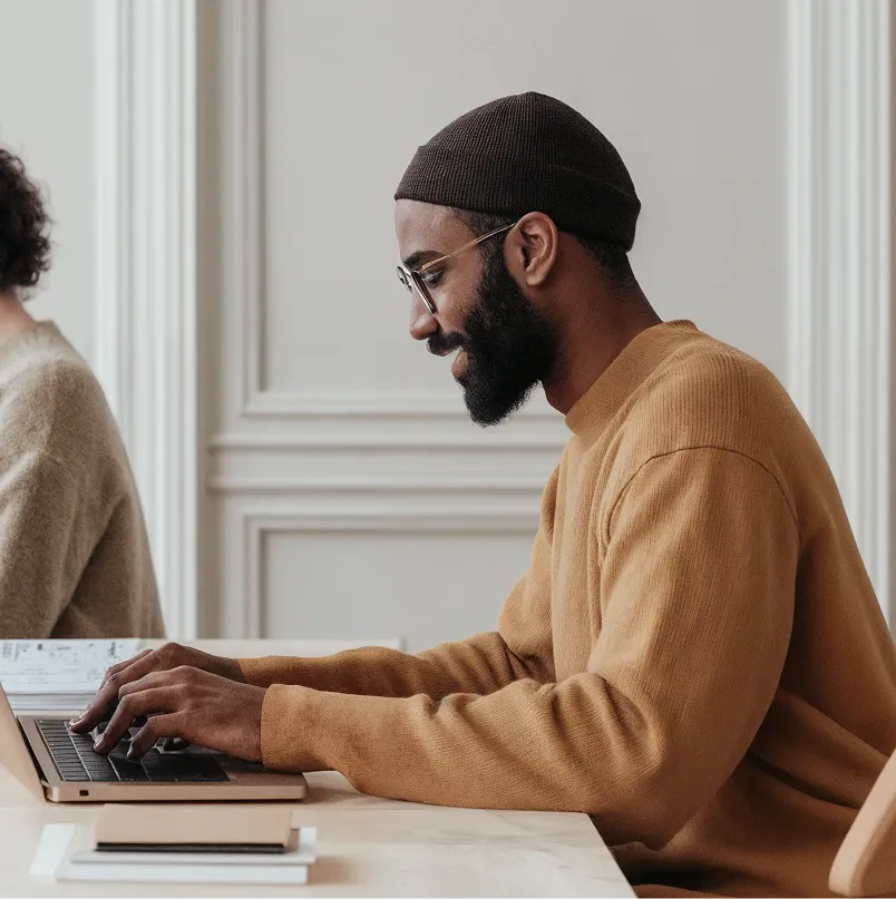 Person wearing glasses and a knit beanie working on a laptop at a desk with notebooks nearby.