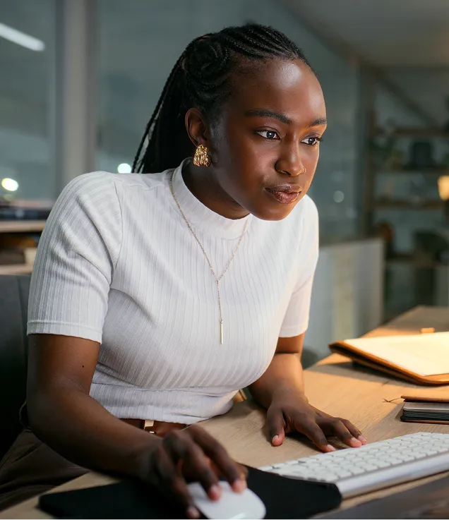 Person seated at a desk, focused on a computer screen while using a mouse and keyboard. The workspace is softly lit, with documents and office items visible on the desk.