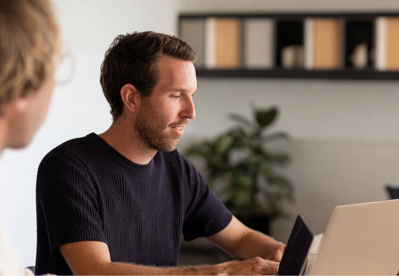 Person sitting at a desk and working on a laptop, viewed in profile, with another person partially visible in the foreground. A shelf with books and a potted plant are visible in the background.