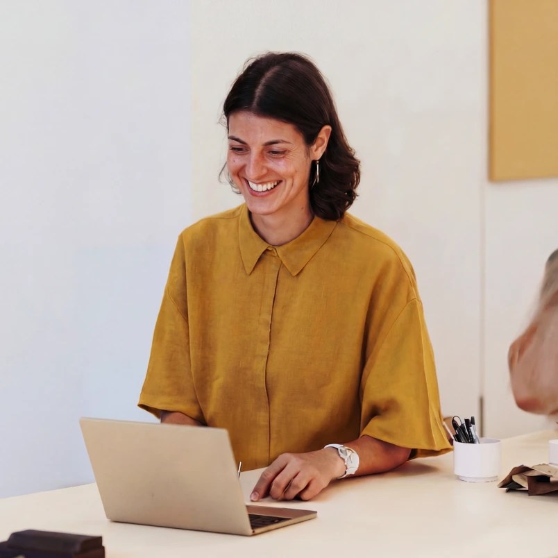 Person in a mustard-colored shirt smiling while working on a laptop at a desk, with a pen holder and another person partially visible beside them.