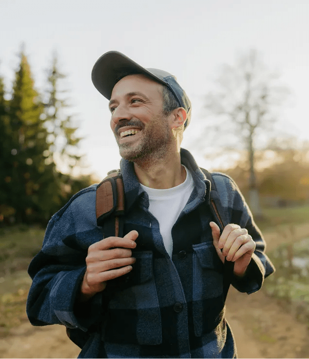 A smiling person wearing a backpack and cap while walking along an outdoor trail surrounded by trees.