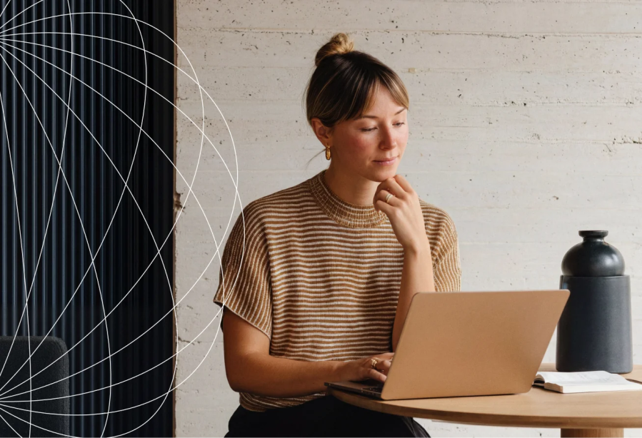 Woman in a striped shirt sits at a table using her laptop, perhaps exploring solutions like WordPress VIP for enterprise brands, with a notebook and vase beside her.